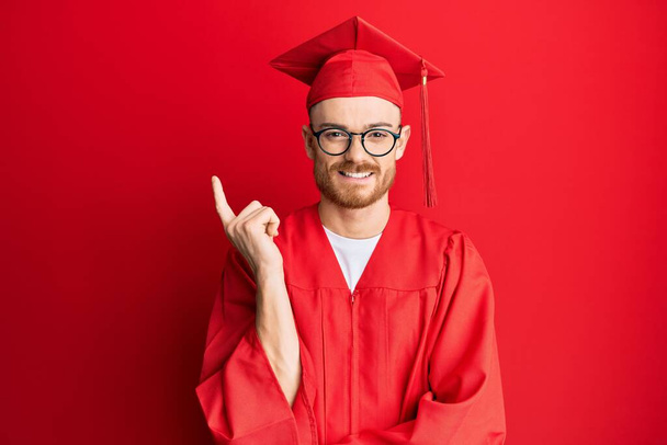 Kırmızı mezuniyet şapkası ve yüzünde kocaman bir gülümseme olan kırmızı saçlı genç adam kameraya el ve parmağıyla bakıyor..  - Fotoğraf, Görsel