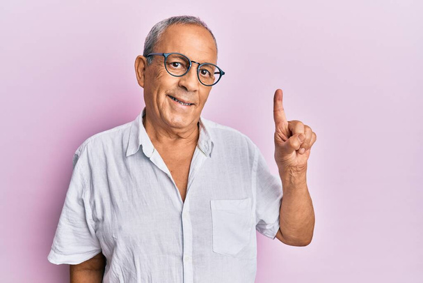 bel homme âgé mature portant chemise décontractée et des lunettes souriant avec une idée ou une question pointant du doigt avec le visage heureux, numéro un  - Photo, image