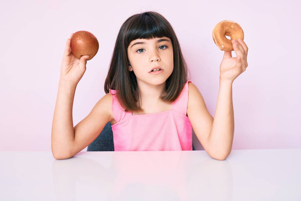 Niña joven con flequillo sosteniendo manzana roja y rosquilla sentada en la mesa expresión despistada y confusa. concepto de duda.  - Foto, Imagen