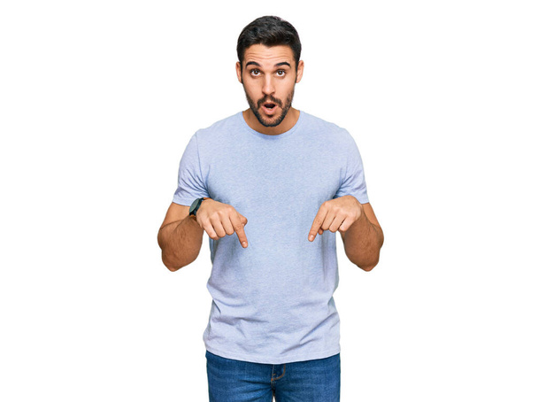 Joven hombre hispano usando ropa casual apuntando hacia abajo con los dedos mostrando publicidad, cara sorprendida y boca abierta  - Foto, Imagen