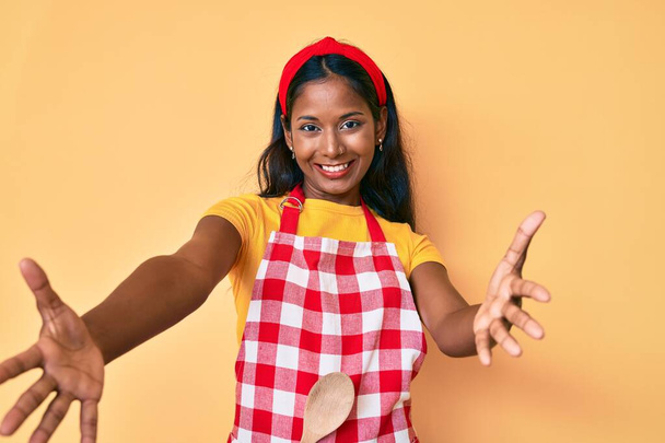 Mladá indiánka v pekařské zástěře se dívá do kamery a usmívá se s otevřenou náručí, aby ji objala. veselý výraz objímající štěstí.  - Fotografie, Obrázek