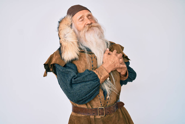 Starý starší muž s šedivými vlasy a dlouhými vousy v tradičním vikingském kostýmu, usmívající se s rukama na hrudi, oči zavřené vděčným gestem na tváři. zdravotní koncepce.  - Fotografie, Obrázek