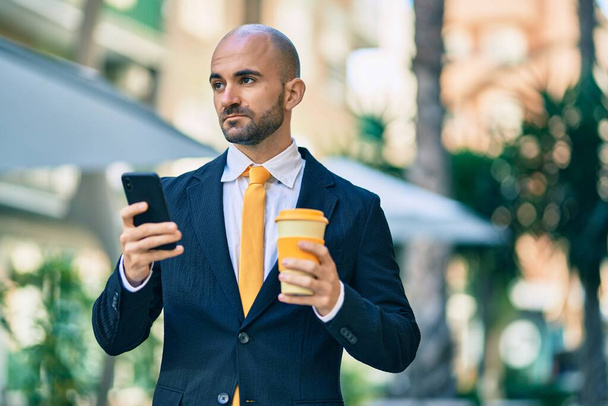 Молодой латиноамериканец лысый бизнесмен с серьезным выражением лица с помощью смартфона пьет кофе в городе. - Фото, изображение