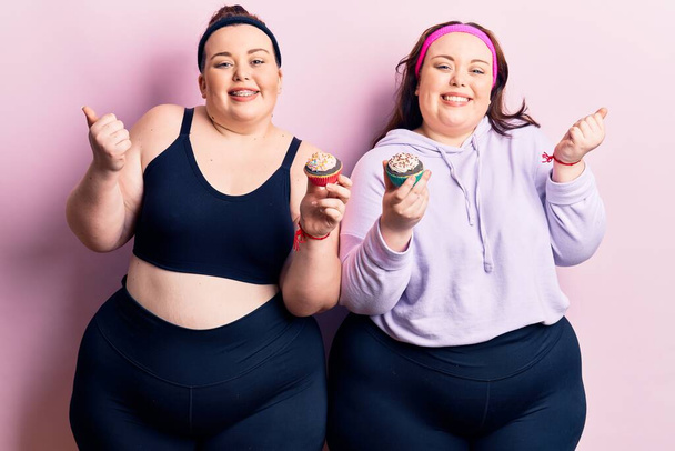 Young plus size twins wearing sportswear holding cupcake screaming proud, celebrating victory and success very excited with raised arm  - Photo, Image