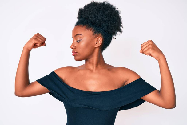 アフリカ系アメリカ人の若い女性が腕の筋肉を示すカジュアルな服を着て誇らしげに笑っている。フィットネスのコンセプト.  - 写真・画像