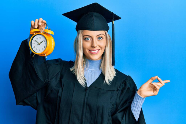 Красива блондинка в шапці для випускних і церемонії одягу тримає будильник усміхнений щасливий вказівник рукою і пальцем на бік
  - Фото, зображення
