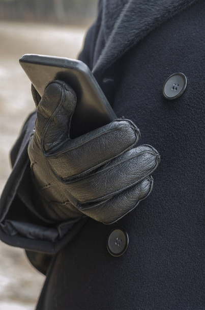 Επιχειρηματίας με μαύρο παλτό και μαύρα δερμάτινα γάντια κρατά το τηλέφωνο στο χέρι του καθώς περπατάει στο πάρκο. Κοντινό πλάνο, επιλεκτική εστίαση. - Φωτογραφία, εικόνα