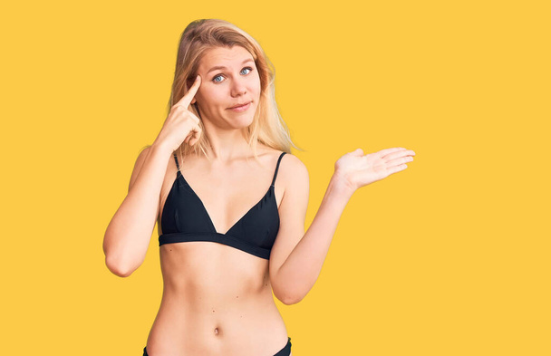 Young beautiful blonde woman wearing bikini confused and annoyed with open palm showing copy space and pointing finger to forehead. think about it.  - Photo, Image