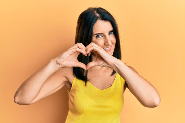 Femme brune d'âge moyen portant des vêtements décontractés sur fond jaune souriant en amour faisant forme de symbole de coeur avec les mains. concept romantique.  - Photo, image