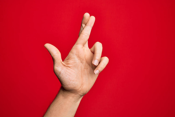孤立した赤い背景のジェスチャーの指の上に指を示す白人の若い男の手が交差,迷信と幸運ジェスチャー,幸運と希望の表現 - 写真・画像