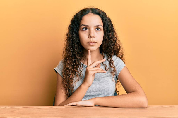 Teenager hispanic girl wearing casual clothes sitting on the table thinking concentrated about doubt with finger on chin and looking up wondering  - Photo, image