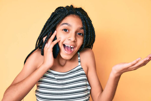 Junge afrikanisch-amerikanische Mädchen mit Zöpfen unterhalten sich auf dem Smartphone und feiern ihre Leistung mit glücklichem Lächeln und Siegerausdruck mit erhobener Hand  - Foto, Bild
