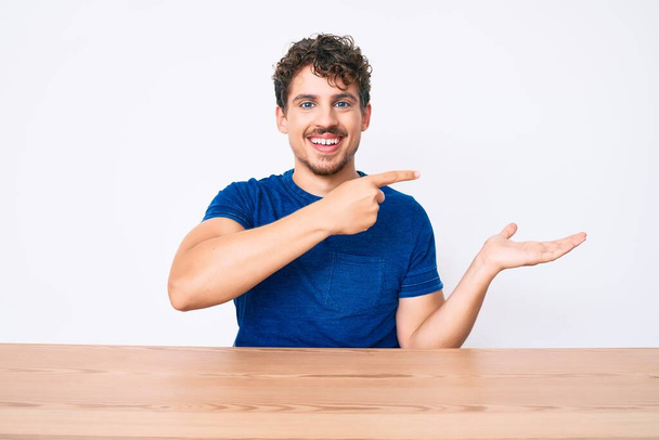 Νεαρός καυκάσιος άνδρας με σγουρά μαλλιά φορώντας καθημερινά ρούχα κάθεται στο τραπέζι έκπληκτος και χαμογελώντας στην κάμερα, ενώ παρουσιάζει με το χέρι και δείχνοντας με το δάχτυλο.  - Φωτογραφία, εικόνα