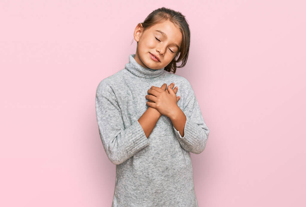 Egy gyönyörű kislány alkalmi garbó pulóvert visel, aki csukott szemmel és hálás gesztussal mosolyog a mellkasára. egészségügyi koncepció.  - Fotó, kép