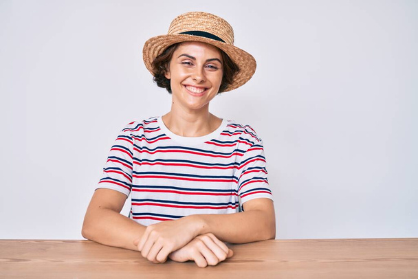 Giovane donna ispanica indossa abiti casual e cappello seduto sul tavolo con un sorriso felice e fresco sul viso. persona fortunata.  - Foto, immagini