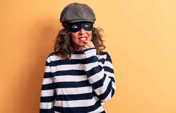 Middle age brunette burglar woman wearing mask and cap standing over yellow background thinking concentrated about doubt with finger on chin and looking up wondering - 写真・画像