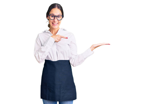 Piękna brunetka młoda kobieta w profesjonalnym fartuchu kelnerka zdumiony i uśmiechnięty do kamery podczas prezentacji z ręki i wskazując palcem.  - Zdjęcie, obraz