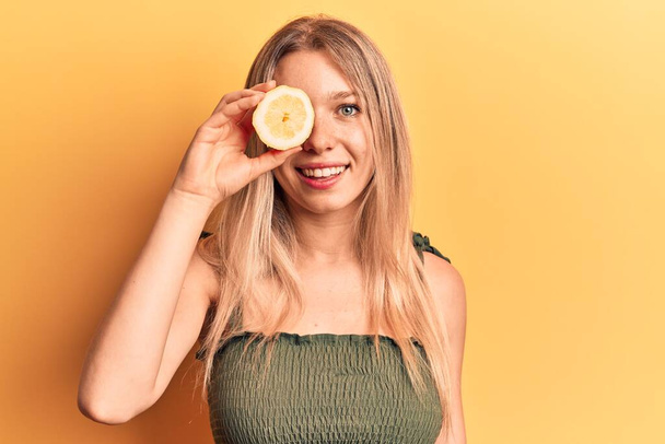Young blonde woman holding lemon looking positive and happy standing and smiling with a confident smile showing teeth  - 写真・画像