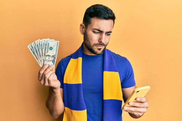 Jeune homme hispanique supporter de football à l'aide d'un smartphone tenant des billets en dollars américains sceptiques et nerveux, fronçant les sourcils bouleversés en raison de problèmes. personne négative.  - Photo, image