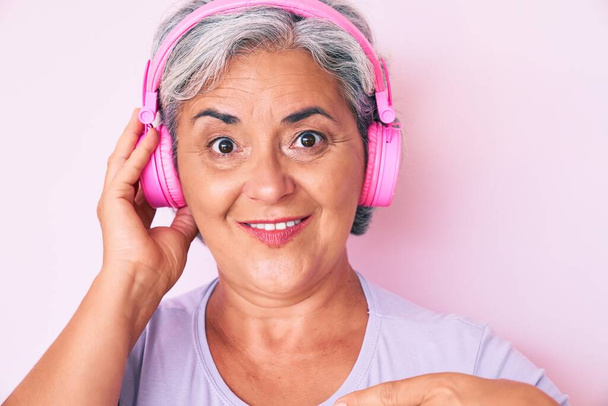 Старшая латиноамериканка слушает музыку, используя наушники, указывая пальцем на себя, улыбаясь счастливой и гордой  - Фото, изображение