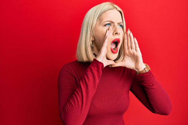 Νεαρή ξανθιά γυναίκα που φοράει καθημερινά ρούχα φωνάζοντας θυμωμένα δυνατά με τα χέρια πάνω από το στόμα  - Φωτογραφία, εικόνα