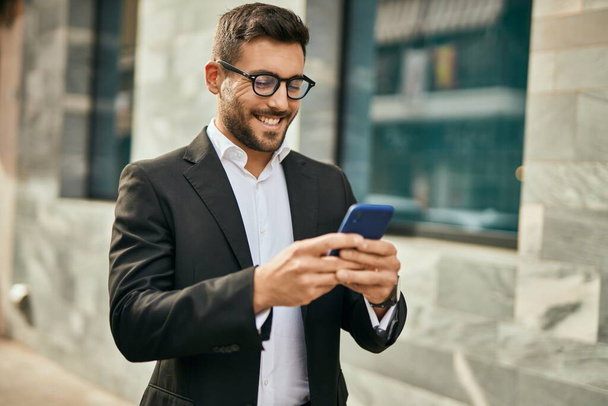 Молодий іспаномовний бізнесмен посміхається щасливим, використовуючи смартфон у місті
. - Фото, зображення
