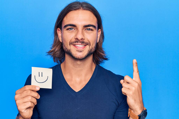 Homem bonito jovem segurando sorriso emoji lembrete sorrindo com uma ideia ou pergunta apontando dedo com rosto feliz, número um  - Foto, Imagem