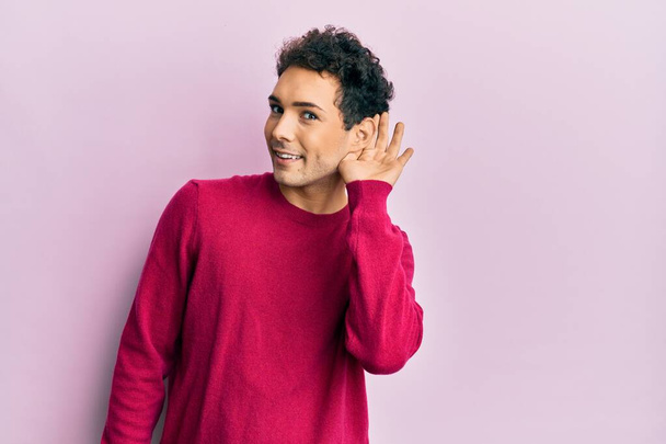 Красивый латиноамериканец в повседневной одежде на розовом фоне улыбается рукой за ухом слушая и слушая слухи или сплетни. концепция глухоты.  - Фото, изображение
