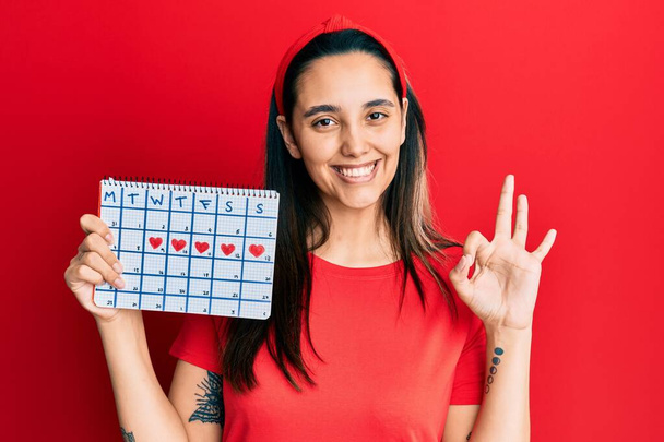 Joven mujer hispana sosteniendo el calendario del corazón haciendo signo bien con los dedos, sonriendo gesto amistoso excelente símbolo  - Foto, Imagen