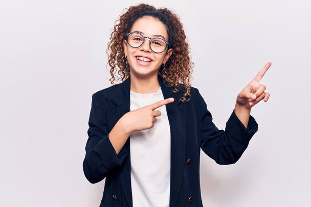 Beautiful kid girl with curly hair wearing business clothes and glasses smiling and looking at the camera pointing with two hands and fingers to the side.  - Photo, Image