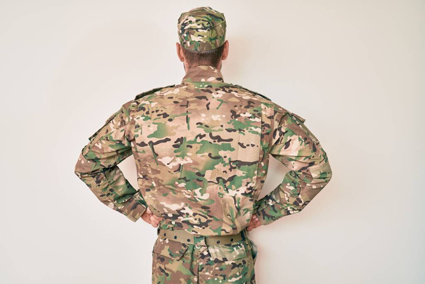 Nuori valkoihoinen mies naamioituneessa armeijapuvussa seisoo selkä menosuuntaan kädet vartalollaan.  - Valokuva, kuva