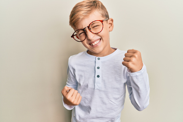 Kleiner kaukasischer Junge in lässiger Kleidung und Brille feiert überrascht und erstaunt den Erfolg mit erhobenen Armen und geschlossenen Augen  - Foto, Bild