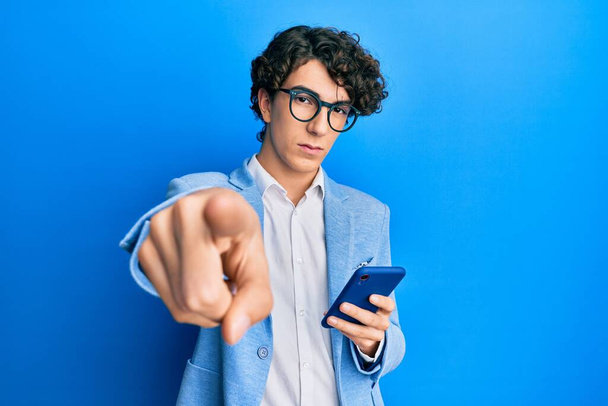 Іспанський молодий чоловік, який користується смартфоном у діловій куртці, вказує пальцем на камеру і на вас, впевнений жест виглядає серйозно  - Фото, зображення
