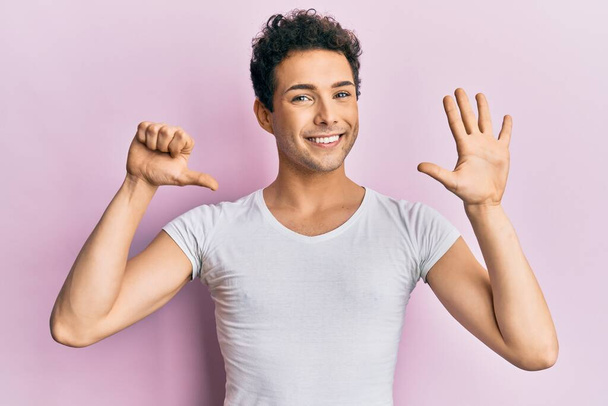 Joven hombre guapo con camiseta blanca casual mostrando y apuntando hacia arriba con los dedos número seis mientras sonríe confiado y feliz.  - Foto, Imagen