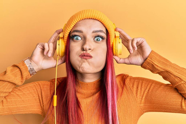 Mladá běloška, která poslouchá hudbu, používá sluchátka, napuchlá tváře s legračním obličejem. ústa nadýchaná vzduchem, lapající vzduch.  - Fotografie, Obrázek