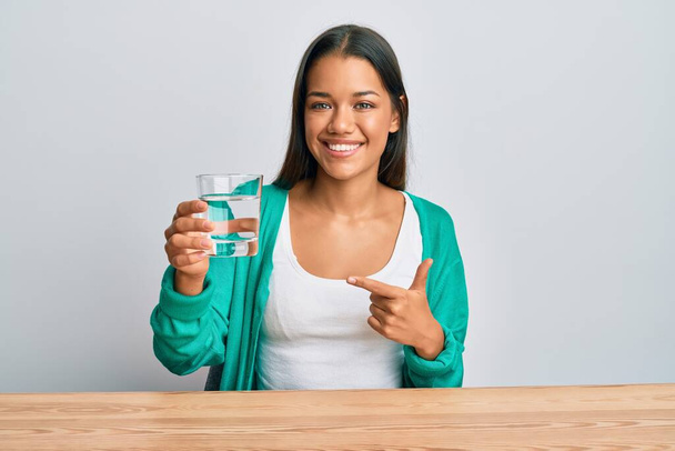 Krásná hispánská žena pije sklenici vody s úsměvem šťastný ukazující rukou a prstem  - Fotografie, Obrázek