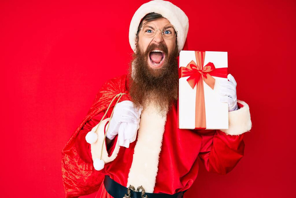 schöner junger roter Mann mit langem Bart im Weihnachtsmann-Kostüm und großer Tasche mit Geschenken, wütend und wahnsinnig schreiend frustriert und wütend, schreiend vor Wut. Wut und aggressives Konzept.  - Foto, Bild