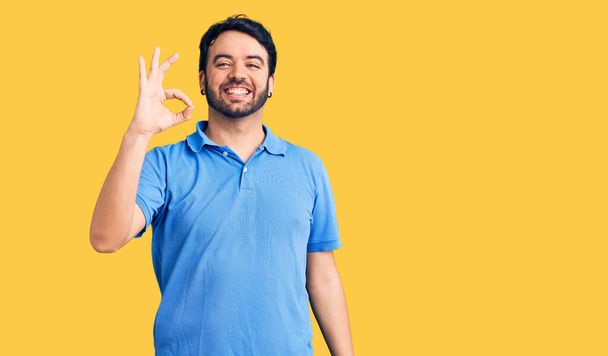 Young hispanic man wearing casual clothes smiling positive doing ok sign with hand and fingers. successful expression.  - 写真・画像
