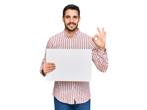 Joven hispano sosteniendo una pancarta vacía en blanco haciendo buen signo con los dedos, sonriendo amistoso gesto excelente símbolo  - Foto, Imagen