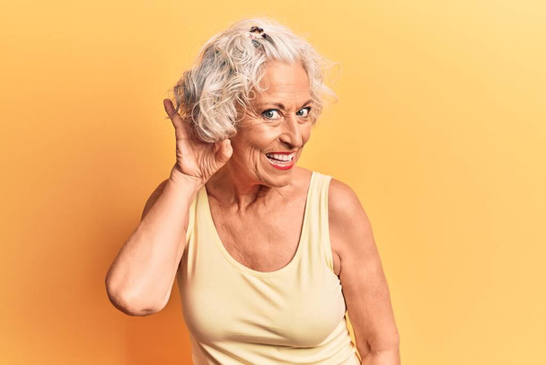 Femme âgée aux cheveux gris portant des vêtements décontractés souriant avec la main sur l'oreille en écoutant une rumeur ou des commérages. concept de surdité.  - Photo, image