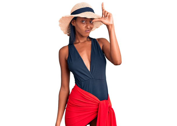 Νεαρή Αφροαμερικανή που φοράει μαγιό και καλοκαιρινό καπέλο κοροϊδεύοντας ανθρώπους με δάχτυλα στο μέτωπο κάνοντας αποτυχημένες χειρονομίες χλευάζοντας και προσβάλλοντας.  - Φωτογραφία, εικόνα