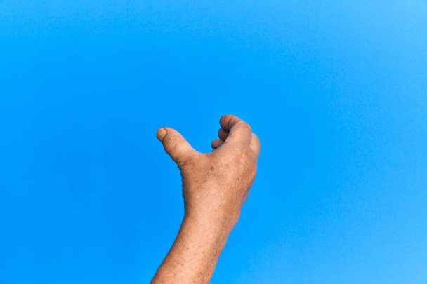 Рука старшего латиноамериканца на синем изолированном фоне с невидимым объектом, пустой рукой, делающей вырезку и хватающий жест  - Фото, изображение