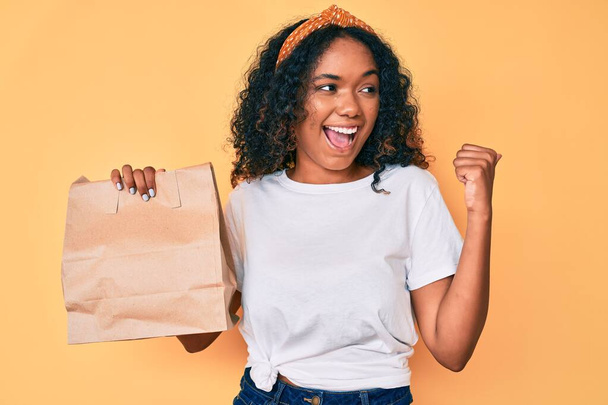Young african american woman holding take away paper bag pointing thumb up to the side smiling happy with open mouth  - Valokuva, kuva