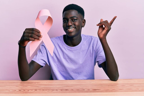 Young african american man holding pink cancer ribbon sitting on the table smiling happy pointing with hand and finger to the side  - Photo, image