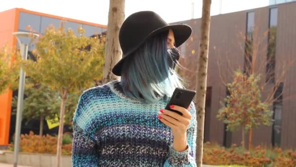 κορίτσι με μπλε μαλλιά σε προστατευτική μαύρη μάσκα και καπέλο με smartphone. Αστική στολή μόδας. - Πλάνα, βίντεο