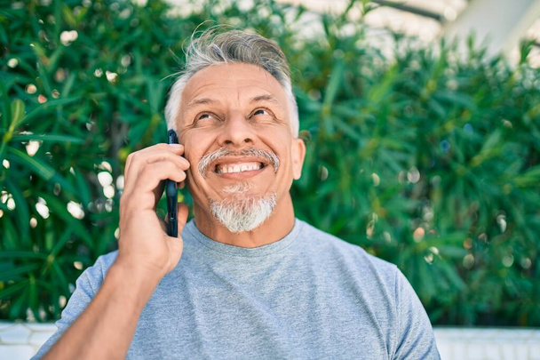 Der grauhaarige Mann mittleren Alters lächelt fröhlich auf dem Smartphone im Park. - Foto, Bild