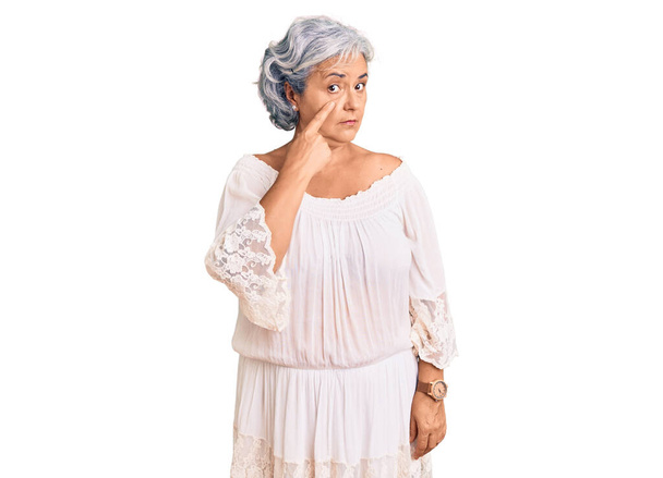 Mujer mayor con el pelo gris usando estilo bohemio apuntando al ojo observándote gesto, expresión sospechosa  - Foto, Imagen