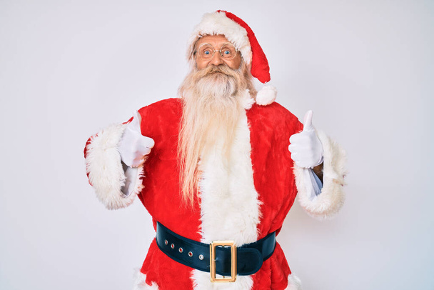 Alter älterer Mann mit grauen Haaren und langem Bart im Weihnachtsmann-Kostüm mit Hosenträgern und positiver Geste mit der Hand, Daumen hoch lächelnd und glücklich. Fröhlicher Ausdruck und Siegergeste.  - Foto, Bild