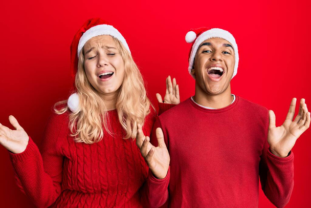Νεαρό διαφυλετικό ζευγάρι φορώντας χριστουγεννιάτικο καπέλο γιορτάζει τρελός και τρελός για την επιτυχία με τα χέρια ψηλά και κλειστά μάτια ουρλιάζοντας ενθουσιασμένος. έννοια νικητή  - Φωτογραφία, εικόνα
