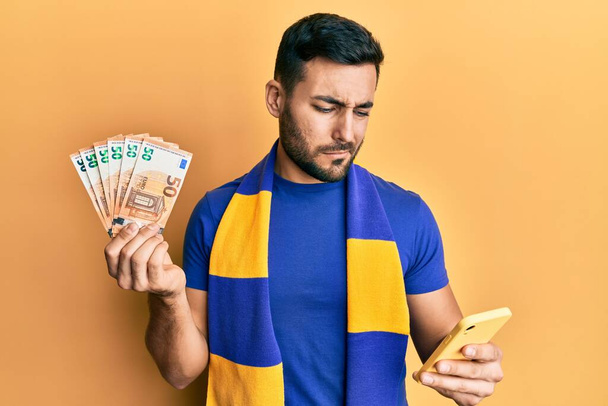 Mladý hispánský fotbalový fanoušek používající smartphone s 50 eurobankovkami, bezradný a zmatený výraz. pochybný koncept.  - Fotografie, Obrázek